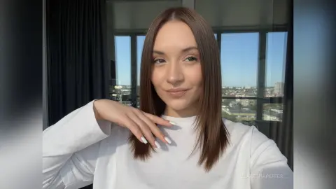Smiling young woman in a white long-sleeve shirt posing by a bright window with a city view.