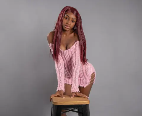 Young woman with long red hair wearing a cozy pink knit sweater sitting on a stool against a gray background.