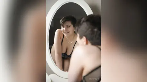Smiling young woman in a black lace bra looking into a round mirror in a bright, minimalist room.