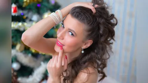 MontseFerrera poses with her hands in her wavy brown hair, wearing red lipstick and pearl earrings against a blurred Christmas tree background.