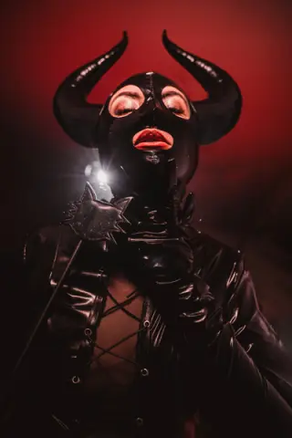 Woman in black latex demon costume with horns and spiked armor against a moody red background.