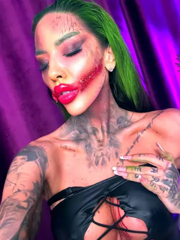 Woman with green hair and horror makeup wearing a black cutout top against a purple backdrop.