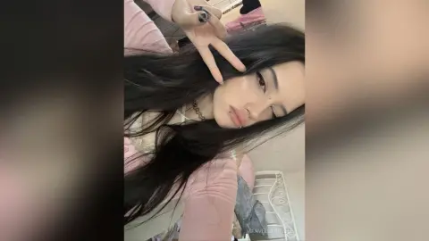MarvellaRovella with long dark hair poses in a pink top and makes a peace sign at the camera.