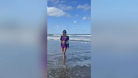 Young woman in purple workout set standing in ocean waves under a blue sky at a sunny beach.