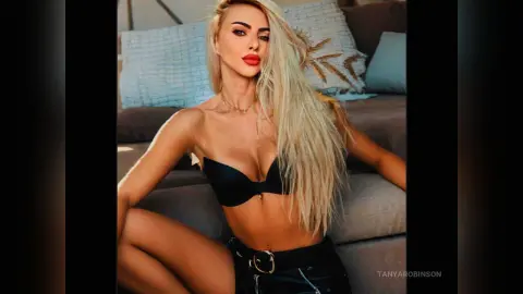 Blonde woman in black bikini top and leather pants posing confidently on a sofa in a bright room.