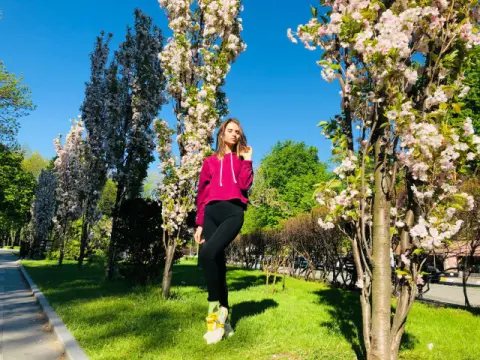 Young woman in a maroon hoodie and black leggings standing by blooming cherry blossom trees on a sunny day.
