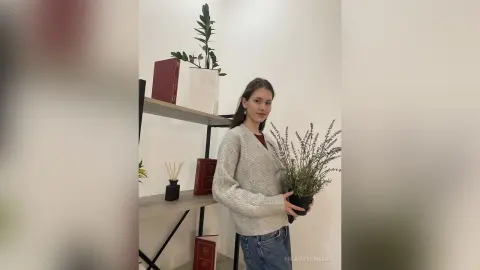 Young woman in a grey knit sweater holding a potted lavender plant in a bright minimalist room.