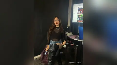 SaraMattheus wearing a black mesh top and denim shorts, sitting in a dark room with a blue dildo on the table next to her.