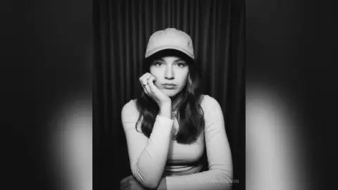 Moody black and white portrait of a young woman in a white long-sleeve shirt and baseball cap against dark curtains.