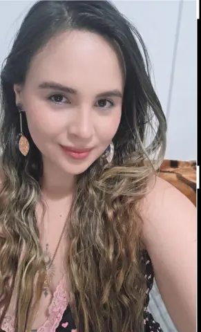Smiling young woman with long wavy hair wearing large bohemian earrings and a lace pink top in a bright room.