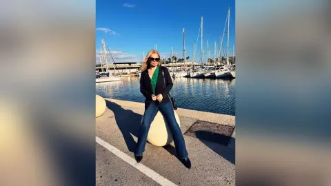 MilenaBia wears sunglasses, a black jacket over a green top, and blue jeans while sitting on a white bollard by a marina filled with sailboats.