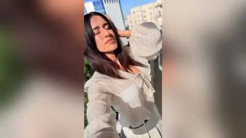 BellaLeen poses with her eyes closed and hands behind her head, wearing a white long-sleeved blouse with a ruffled neckline and long dark hair.