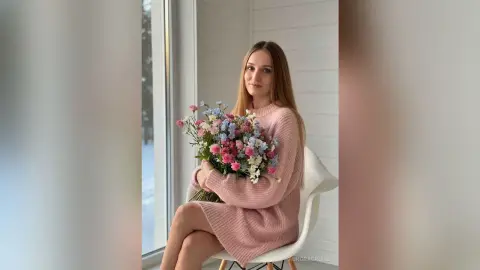 AuroraCruise sits by a window wearing a pink knit sweater while holding a large bouquet of pink and blue flowers.