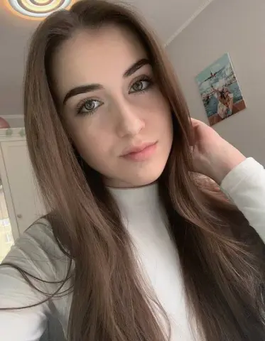 SashaShin poses with her long brown hair, wearing a white turtleneck top against a plain wall background.