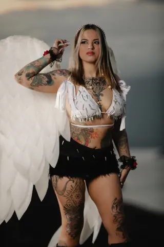 Tattooed woman in white angel wings and a fringed crop top posing outdoors in a dreamy, ethereal mood.
