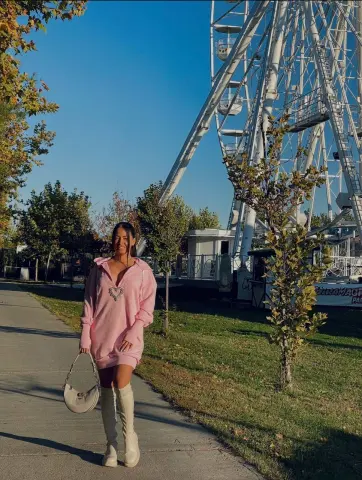 Woman in a pink oversized sweatshirt and white boots walking near a white Ferris wheel on a sunny day.