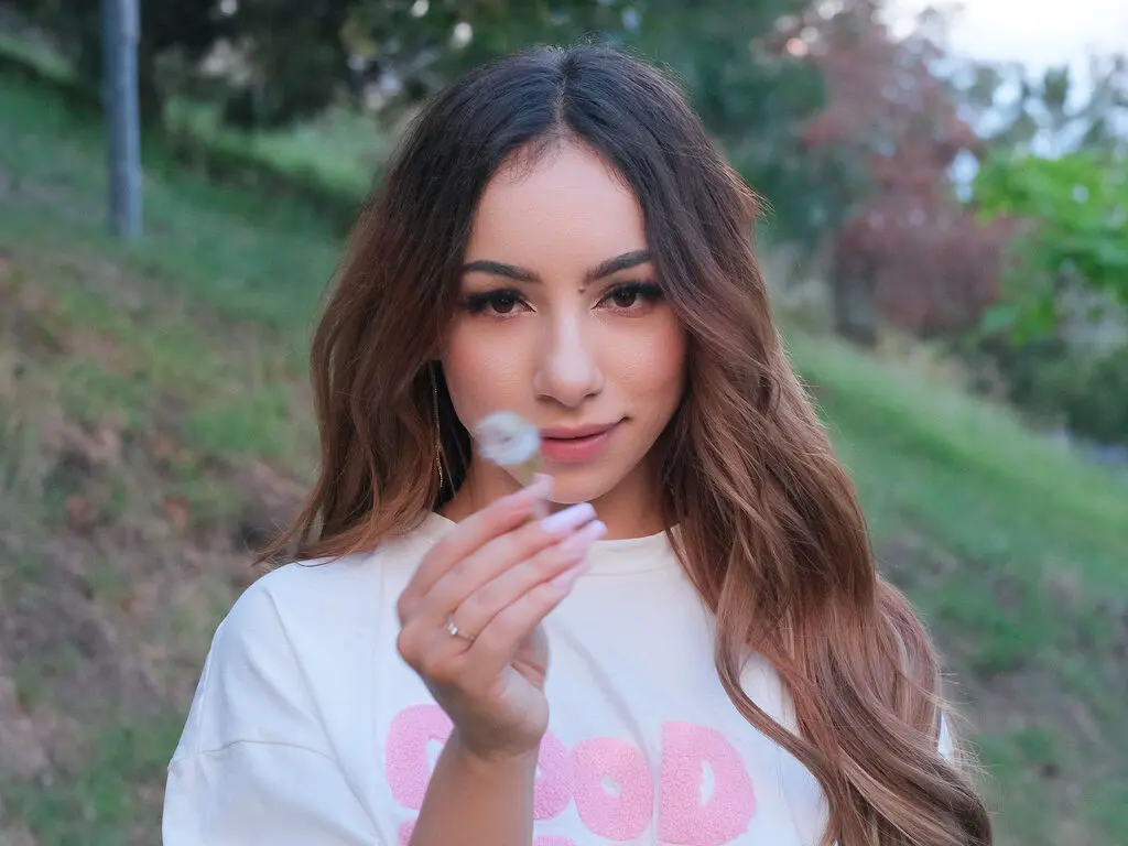 Brunette woman in a white graphic t-shirt holding a dandelion in a soft, dreamy outdoor garden setting.