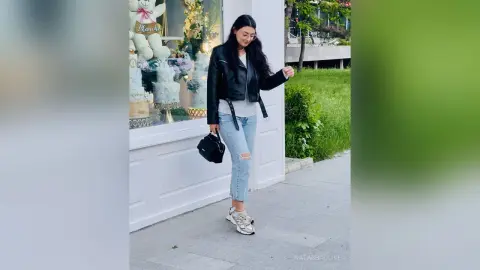 Stylish woman in a black leather jacket and light blue jeans walking past a bakery storefront on a sunny day.