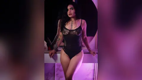Young woman with tattoos in a black lace bodysuit posing in moody purple lighting.