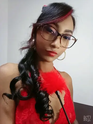 Glamorous woman with long wavy dark hair wearing a red lace dress and glasses in a bright indoor setting.
