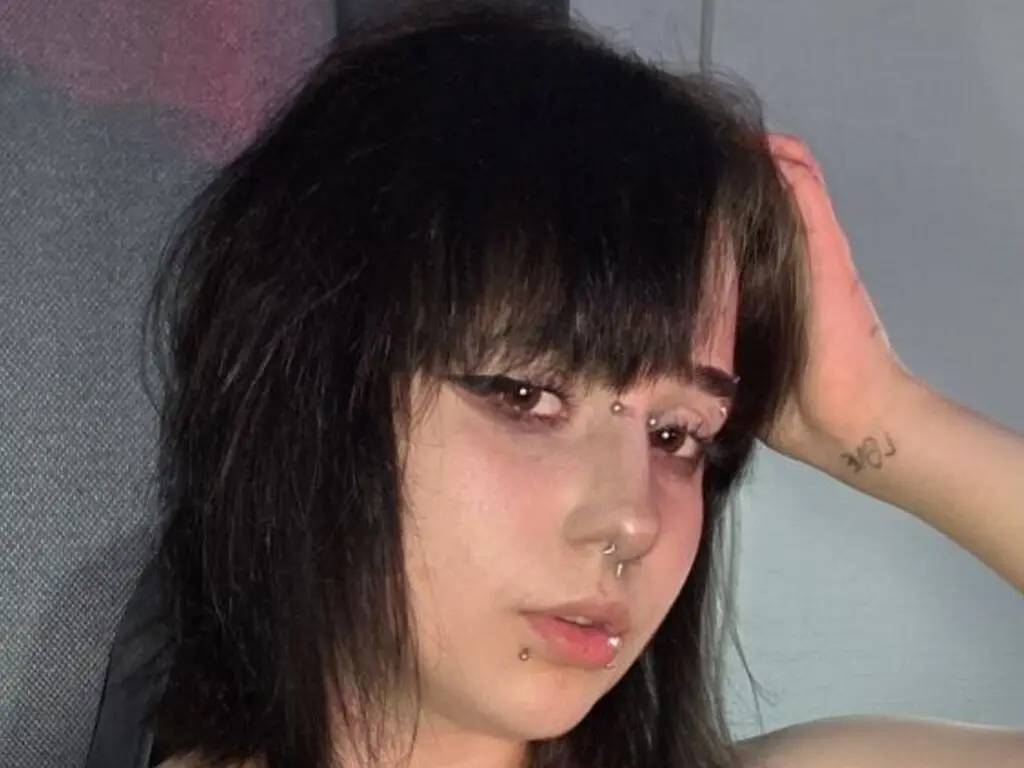 Young woman with dark messy hair and facial piercings posing in a moody indoor setting.