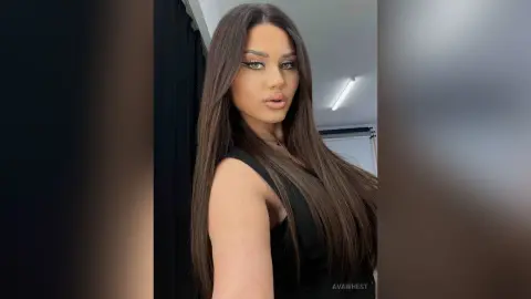 Glamorous woman with long brown hair wearing a black sleeveless top in a modern indoor setting.