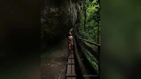 ElizabethSwith stands naked on a wooden walkway next to a large rock wall in a lush, green forest setting.