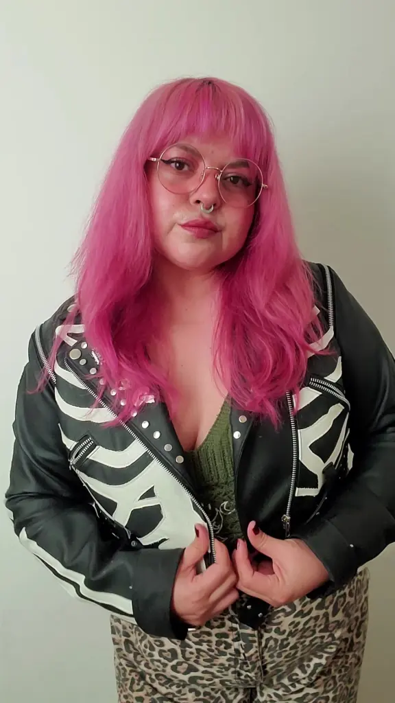 Satyndoll poses with pink hair and red lipstick, wearing a black lace top and a black choker against a purple geometric wall.