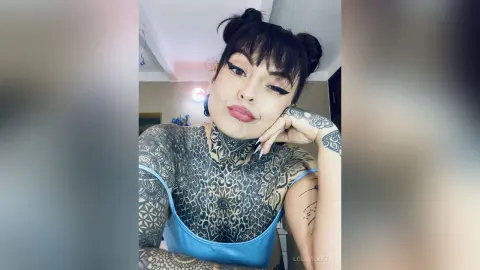 Tattooed woman in a blue camisole posing with a soft expression in a bright indoor setting.