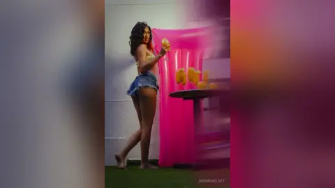 Stylish woman in denim shorts holding a cocktail at a vibrant pink summer party setting.