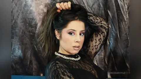Glamorous woman in a black lace long-sleeve top posing against a textured grey backdrop with elegant makeup.