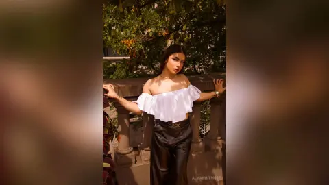 AlmaWhitmore poses outdoors in a white off-the-shoulder ruffled top and black leather pants, leaning against a stone balcony railing.