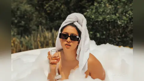 KimRoberth relaxes in a white bubble bath while wearing a white towel wrap on her head and black sunglasses, holding a glass of white wine.