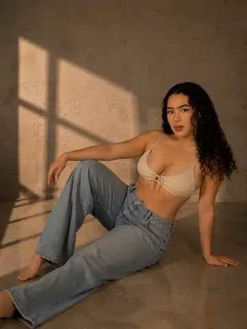 paulinaa_rosee poses sitting on the floor wearing a white bikini top and light blue jeans, showing her long dark curly hair.