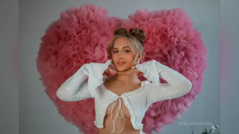 StellaFletcher poses with her hands under her chin against a large pink heart-shaped ruffly background, wearing a white long-sleeved cropped top.
