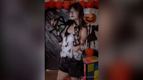 Woman in spooky spider costume and glasses posing in a dark Halloween setting with pumpkin decorations.