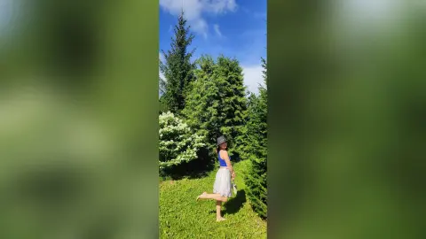 Cheerful woman in a blue tank top and white skirt posing in a lush green summer garden under a blue sky.