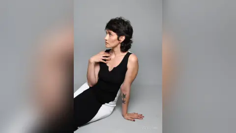 IveteDurand poses in a black and white dress, looking away with her hand resting on her neck against a plain grey background.