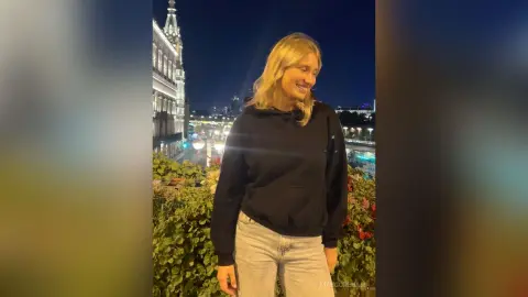 Smiling blonde woman in a black hoodie and light jeans standing on a balcony at night with city lights.