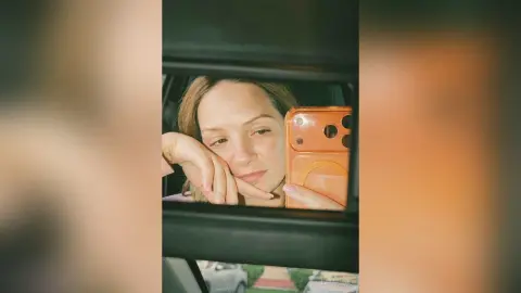 Young woman taking a moody selfie in a car window with an orange phone case.