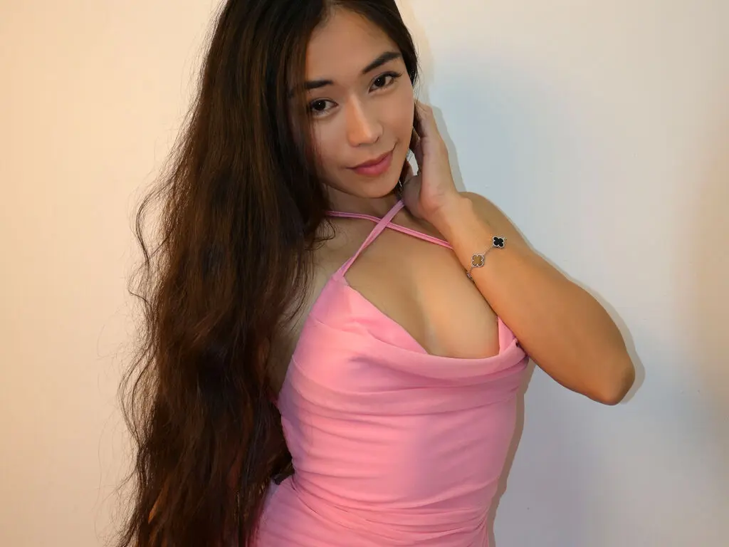 Smiling young woman with long brown hair wearing a pink halter neck dress against a white wall.