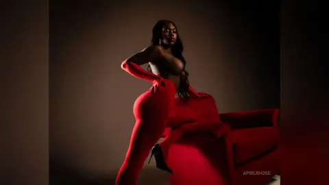 Elegant Black woman in red jumpsuit posing on a red velvet chair against a dark moody background.