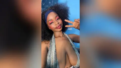 Smiling woman with curly hair and pink eye makeup wearing a silver halter top in a bright blue room.