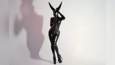 Woman in a sleek black latex bodysuit and horned mask posing against a white studio background.