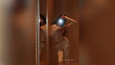 OliviaHardies poses in a white long-sleeve top and white pants, taking a mirror selfie in a dimly lit room.