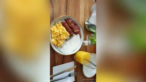 NohomiCambel's breakfast plate features scrambled eggs, sausages, bacon, and a white tortilla on a wooden table.
