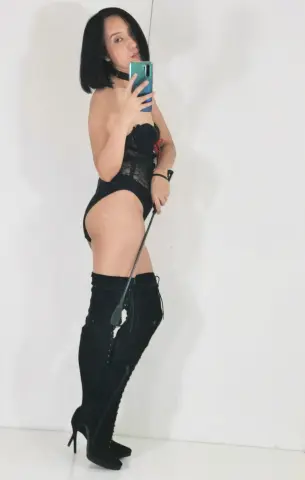 Woman in black lace lingerie and over-the-knee boots taking a mirror selfie in a bright room.