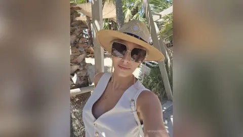 Smiling woman in a white sleeveless top and straw sun hat enjoying a sunny summer day at a tropical beach resort.