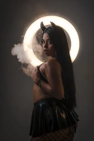 Woman in black devil horns and leather skirt posing amidst mystical smoke and a glowing halo light.