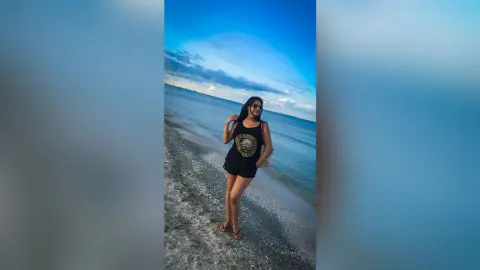 MissyJolie stands on a sandy beach wearing a black tank top and sunglasses while looking toward the camera.
